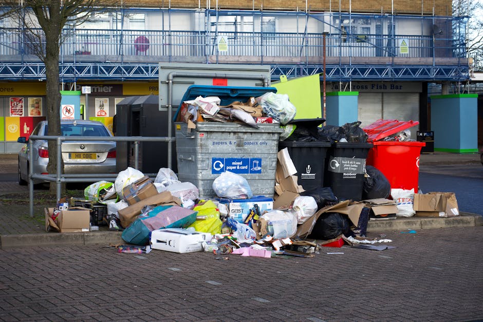 A collection of overflowing rubbish bins and scattered waste bags on a paved sidewalk in an urban area. The central bin is a large, gray, wheeled container labeled for mixed paper and cardboard, with its bright blue lid open and various paper waste protruding. To its right are several black waste bags and a red recycling bin, both filled and overflowing. On the left side of the image, smaller cardboard boxes and plastic packaging are stacked or lying on the ground, along with crumpled paper and assorted packaging materials. Behind the waste area, a metal safety railing separates the rubbish from parked vehicles, including a silver car with a visible yellow and black UK registration plate. In the background, a building with a blue scaffolding structure and partially visible storefronts, along with structural poles, indicates a commercial or retail environment. The scene is lit by natural daylight, revealing the textures and colors of the waste and surroundings, illustrating an example of private rubbish storage likely related to alternative waste handling services such as those provided by Rubbish Collection Fulham.