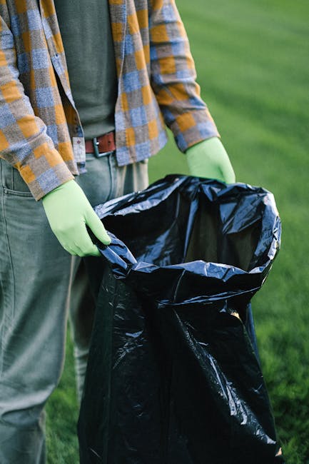 A person wearing green gloves and casual clothing, including a plaid shirt over a green t-shirt and beige trousers, is holding open a large black rubbish bag outdoors on a grassy area. The bag appears to be made of durable plastic, with a shiny, slightly crinkled surface, and its interior is visible as the person prepares to discard waste. The background shows a well-maintained lawn with vibrant green grass, and the natural lighting suggests a daytime setting. This scene reflects an on-site waste collection or rubbish removal activity, which could be associated with private rubbish clearance services offered by Rubbish Collection Fulham. The focus on the bag and gloves highlights the environment-friendly approach and the importance of proper waste handling during rubbish removal processes.