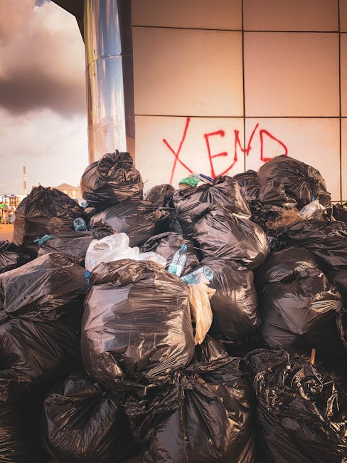 A collection of overflowing rubbish bins and scattered waste bags on a paved sidewalk in an urban area. The central bin is a large, gray, wheeled container labeled for mixed paper and cardboard, with its bright blue lid open and various paper waste protruding. To its right are several black waste bags and a red recycling bin, both filled and overflowing. On the left side of the image, smaller cardboard boxes and plastic packaging are stacked or lying on the ground, along with crumpled paper and assorted packaging materials. Behind the waste area, a metal safety railing separates the rubbish from parked vehicles, including a silver car with a visible yellow and black UK registration plate. In the background, a building with a blue scaffolding structure and partially visible storefronts, along with structural poles, indicates a commercial or retail environment. The scene is lit by natural daylight, revealing the textures and colors of the waste and surroundings, illustrating an example of private rubbish storage likely related to alternative waste handling services such as those provided by Rubbish Collection Fulham.
