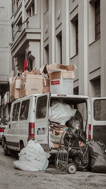 The image shows the open rear doors of a white van parked on a city street, with a large load of discarded rubbish and moving boxes partially visible inside and spilling onto the pavement. Cardboard boxes, some flattened and others still intact, are stacked at the back of the vehicle, with a mix of paper, plastic bags, and packaging materials visible. Several black and white trash bags are also seen, some placed on the ground beside the van, contributing to a cluttered appearance. The scene is set against the backdrop of a multi-storey building with beige or light grey stone or concrete facades, featuring rectangular windows and modern architectural details. Environmental lighting appears natural, suggesting daytime, with shadows cast on the building's surface. This scene reflects private rubbish clearance or on-site waste collection, highlighting the typical contents involved in domestic or commercial clearance within an urban environment. Rubbish Collection Fulham often handles similar debris to facilitate non-local authority waste disposal and alternative waste handling services in Fulham.