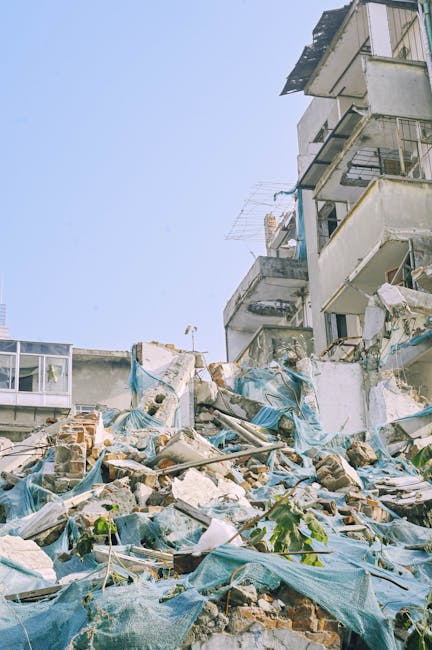 A scene of damage and debris at the base of a multi-storey residential building, with broken bricks, concrete fragments, and scattered building materials. Blue tarpaulins are draped over parts of the rubble, some secured with bricks and debris. The building walls and balconies above show signs of structural damage, with some railings and fixtures leaning or partially dislodged. Windows are visible on the higher floors, with some open and others reflecting sunlight. The background features a clear, pale blue sky. The scene likely results from a recent collapse or demolition, illustrating the need for professional rubbish removal services such as those provided by Rubbish Collection Fulham, who specialise in alternative waste handling and on-site clearance for complex rubbish disposal tasks.
