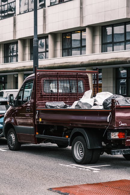 A dark red flatbed utility truck parked on a city street in front of a modern building with large glass windows and a curved concrete facade. The truck's bed is filled with various black and grey rubbish bags and loose debris, likely waste awaiting collection. The truck features a protective metal grille behind the cab, and its rear tailgate is closed, with visible tail lights and wheel arches. The surrounding environment includes a lamppost positioned behind the truck, and adjacent vehicles are partially visible along the street. The scene suggests an area where private waste collection services, such as those provided by companies like Rubbish Collection Fulham, are being used for on-site rubbish removal, offering an alternative to local authority collection. The overall atmosphere is typical of urban rubbish disposal and collection operations, emphasizing the practical handling of waste in a professional manner.