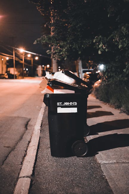 A black wheeled rubbish bin with the label 'St. John's' positioned on a residential pavement at night, with the lid open revealing a partially visible pile of mixed waste, including cardboard boxes, plastic bags, and paper. The bin's surface has a matte finish, and it stands upright on the concrete edge of the pavement, casting a shadow to the right. In the background, there are streetlights illuminating an empty road with a few parked cars and tree-lined sidewalks, creating a dim, urban nighttime scene. The surrounding environment suggests an area where residential waste collection or private rubbish disposal services might operate, supporting the context of alternative waste handling. The scene appears calm and quiet, typical of late evening or early night hours, with soft lighting accentuating the details of the bin and nearby foliage, consistent with professional rubbish removal practices in the Fulham area where Rubbish Collection Fulham provides such services.