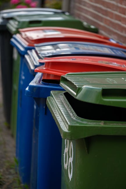 A row of large waste bins lined up along a pavement adjacent to a brick wall, with the front green bin in clear focus displaying a textured plastic lid with horizontal ridges. Behind it, several blue bins with matching textured lids are visible, followed by red bins that also have similar ridged lids, all arranged closely together in a neat, straight formation. The bins are outdoor and likely placed for collection, with some visible graffiti on the green bin's side. In the background, muted greenery and the edge of the brick wall are slightly blurred, with natural daylight illuminating the scene evenly. This arrangement typifies private waste collection options, often provided by local rubbish removal services like Rubbish Collection Fulham, catering to alternative waste handling needs outside of municipal services.