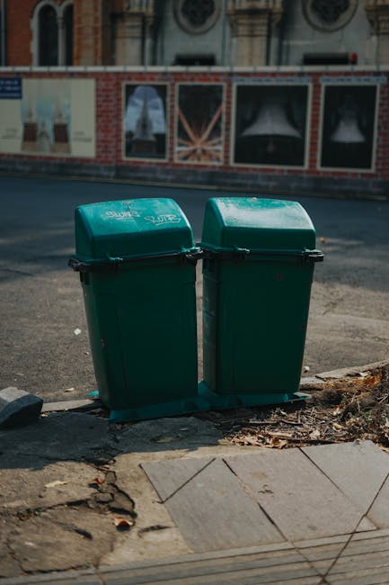 A black wheeled rubbish bin with the label 'St. John's' positioned on a residential pavement at night, with the lid open revealing a partially visible pile of mixed waste, including cardboard boxes, plastic bags, and paper. The bin's surface has a matte finish, and it stands upright on the concrete edge of the pavement, casting a shadow to the right. In the background, there are streetlights illuminating an empty road with a few parked cars and tree-lined sidewalks, creating a dim, urban nighttime scene. The surrounding environment suggests an area where residential waste collection or private rubbish disposal services might operate, supporting the context of alternative waste handling. The scene appears calm and quiet, typical of late evening or early night hours, with soft lighting accentuating the details of the bin and nearby foliage, consistent with professional rubbish removal practices in the Fulham area where Rubbish Collection Fulham provides such services.