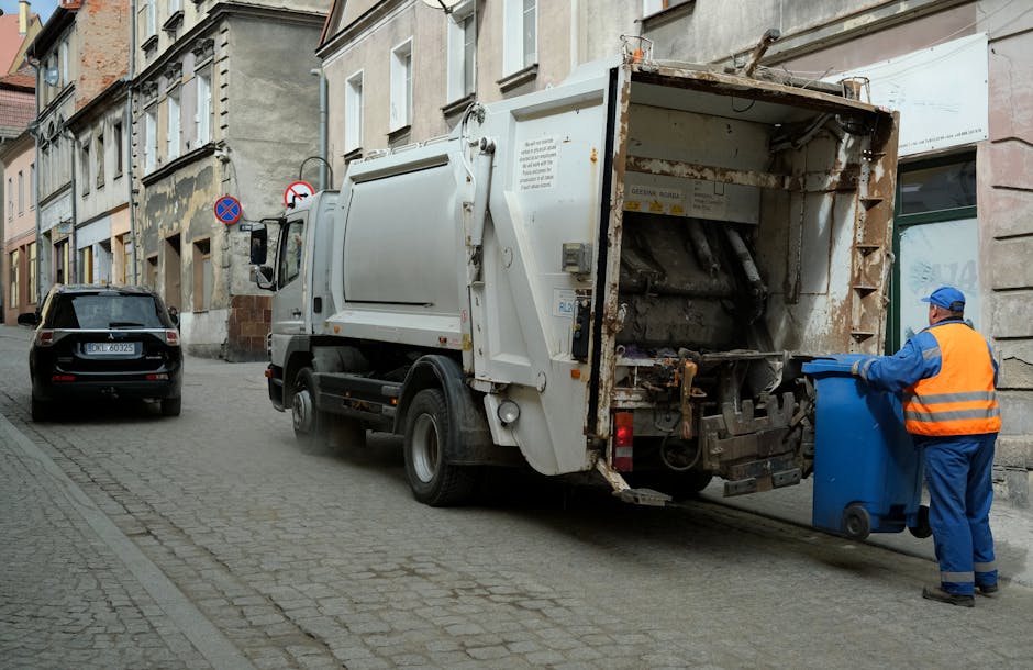 A waste collection truck operated by Rubbish Collection Fulham is parked along a cobblestone street, with the rear hatch open revealing a compartment designed for the disposal of household or commercial rubbish. The truck is white with visible signs of rust and wear on the reinforced steel surface, indicating regular use for waste removal services. A worker dressed in a blue uniform and orange high-visibility vest is standing next to a blue wheeled rubbish bin, actively loading it into the rear compartment of the truck. The street is lined with older, multi-story buildings featuring mixed materials such as bricks and plaster, with some weathering and peeling paint visible on the facades. In the background, parked cars, including a black vehicle, are stationary along the curb, with street signs visible on the building walls. The natural ambient lighting suggests a cloudy day, and the scene highlights an example of private waste collection or on-site rubbish removal typically carried out by independent waste disposal services in urban environments.