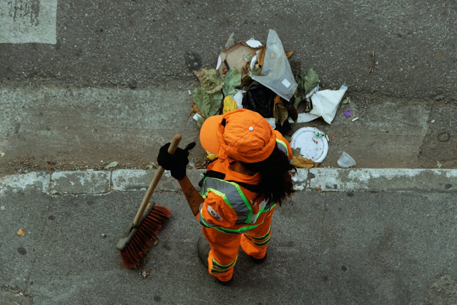 An individual dressed in high-visibility orange work clothing, including a matching hat and reflective safety vest, is seen from above, sweeping a paved sidewalk with a long-handled brown broom. The person is wearing black gloves and is positioned near the edge of the pavement, close to a weathered concrete wall that separates the sidewalk from a small pile of mixed waste. The rubbish consists of various materials such as crumpled paper, leaves, plastic wrappers, and a discarded white paper plate, with some trash partially spilling onto the sidewalk. The scene is outdoors and appears to be part of a street cleaning or rubbish collection effort, with the surrounding environment including a rough cement surface and a narrow strip of dirt or debris along the wall. The lighting is natural, suggesting daytime conditions. This image illustrates the type of on-site clearance services provided by companies like Rubbish Collection Fulham, especially for private waste collection and rubbish removal tasks in urban areas.