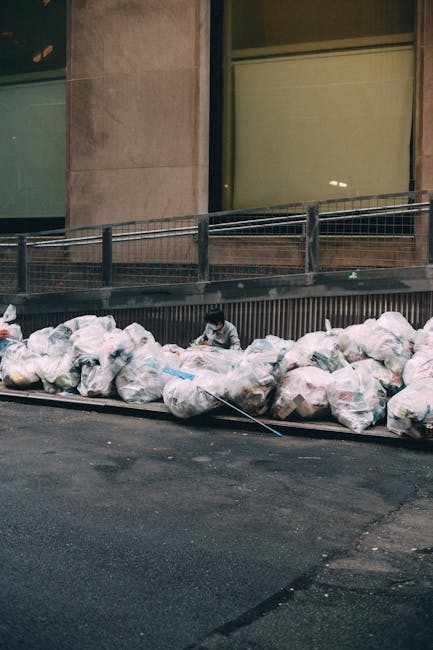 A waste collection truck operated by Rubbish Collection Fulham is parked along a cobblestone street, with the rear hatch open revealing a compartment designed for the disposal of household or commercial rubbish. The truck is white with visible signs of rust and wear on the reinforced steel surface, indicating regular use for waste removal services. A worker dressed in a blue uniform and orange high-visibility vest is standing next to a blue wheeled rubbish bin, actively loading it into the rear compartment of the truck. The street is lined with older, multi-story buildings featuring mixed materials such as bricks and plaster, with some weathering and peeling paint visible on the facades. In the background, parked cars, including a black vehicle, are stationary along the curb, with street signs visible on the building walls. The natural ambient lighting suggests a cloudy day, and the scene highlights an example of private waste collection or on-site rubbish removal typically carried out by independent waste disposal services in urban environments.