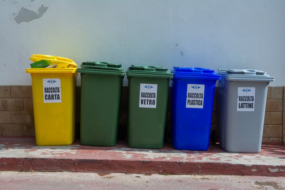 A row of five plastic waste bins is positioned against an external wall with a brick base and a plain, light-colored upper surface. The bins are arranged on a paved surface with a slightly raised curb at the front. From left to right, the bins are yellow, green, darker green, blue, and grey. Each bin has a hinged lid and is labeled with white stickers featuring black text and an eye logo, indicating different types of recyclable waste: 'RACCOLTA CARTA' for paper, 'RACCOLTA VETRO' for glass, 'RACCOLTA PLASTICA' for plastic, and 'RACCOLTA LATTINE' for cans. The yellow bin on the far left contains smaller yellow waste items, possibly lids or containers, and the lids are slightly ajar. The environment appears to be an outdoor area possibly near a residence or commercial premises, with natural lighting illuminating the scene. The image relates to waste collection services, illustrating the separation of recyclables as part of an independent waste disposal approach.